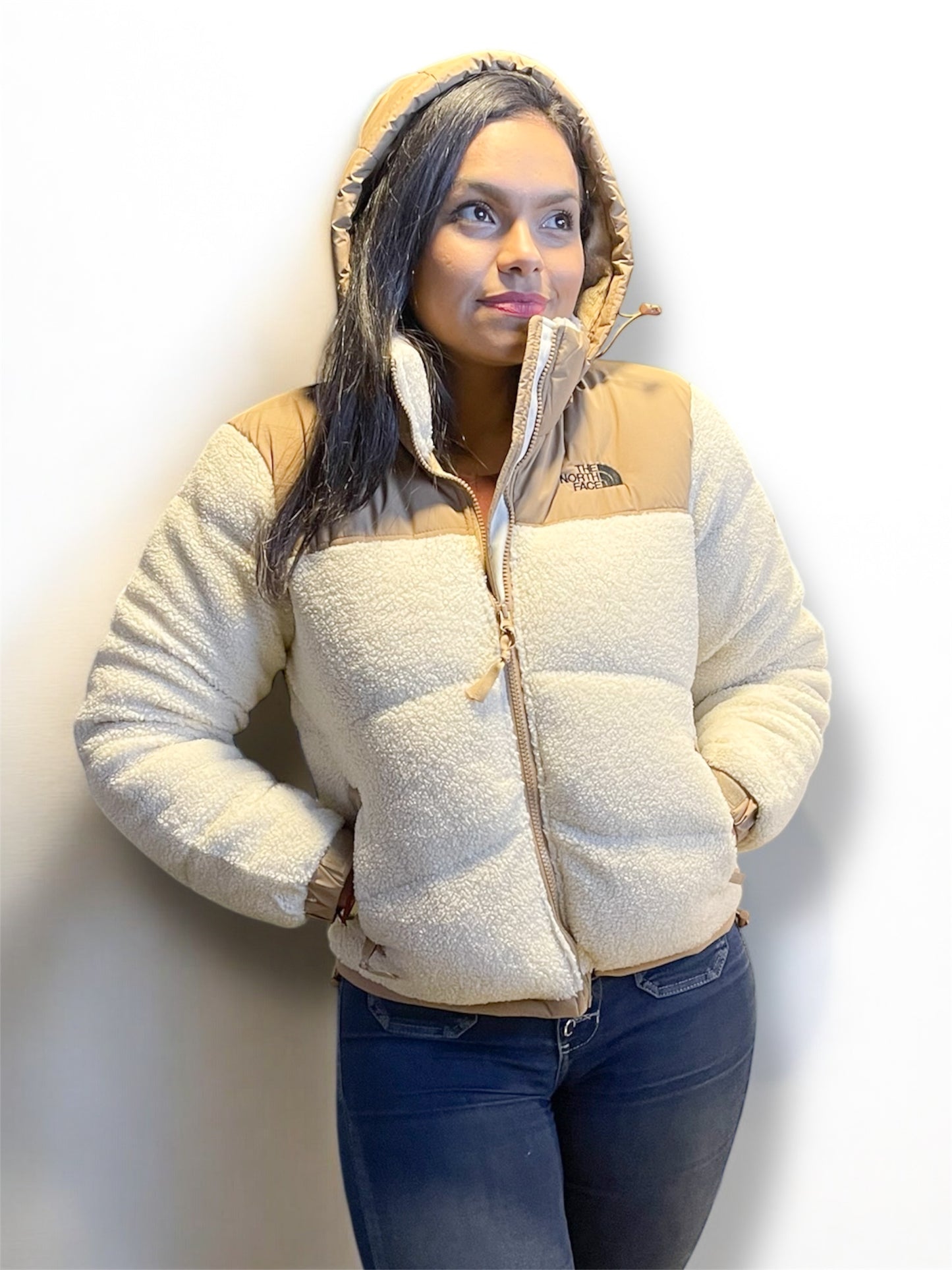 Mujer usando chaqueta teddy beige con capucha, manos en los bolsillos, fondo blanco.