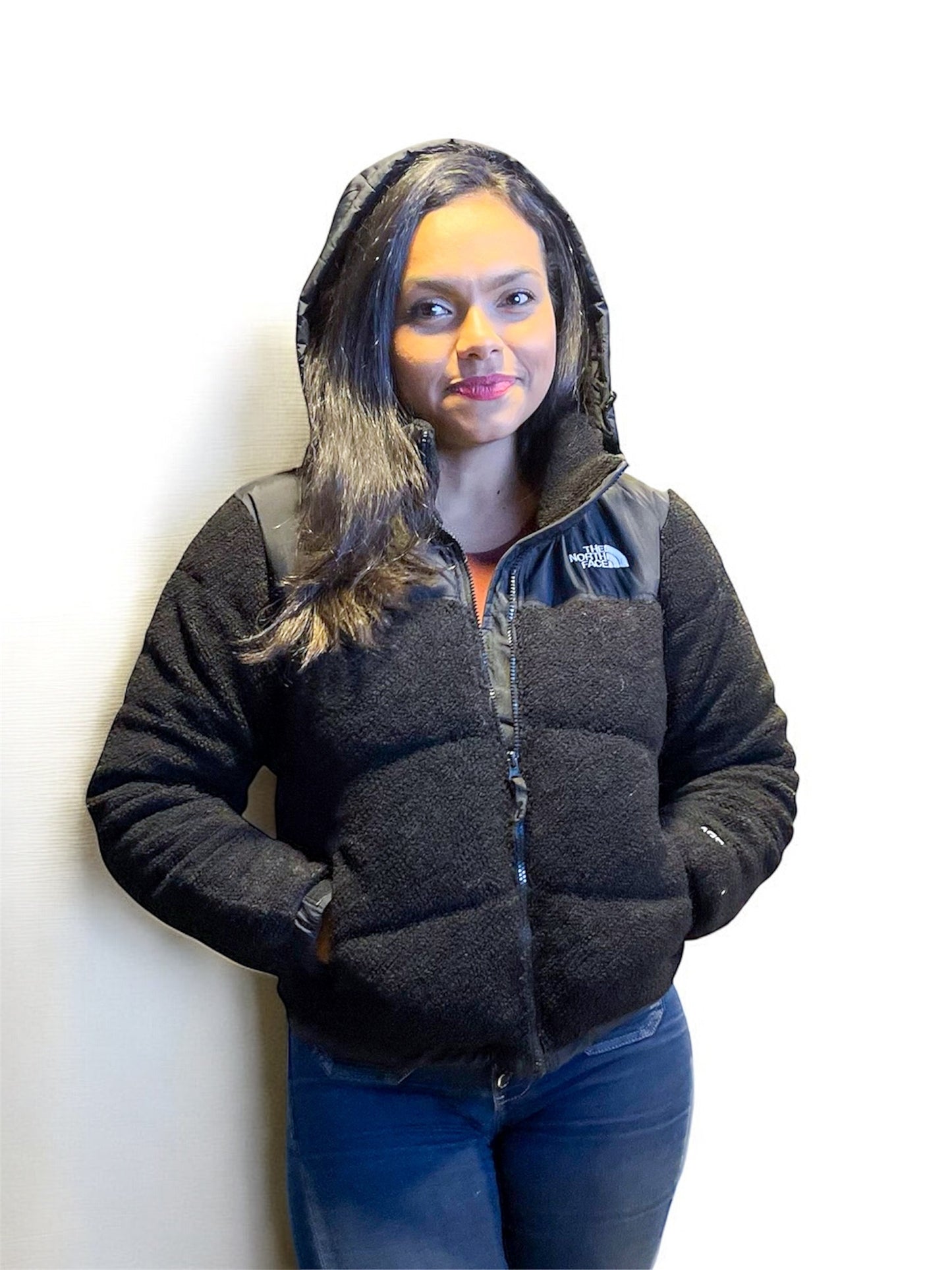Mujer con chaqueta tipo borrego negra con capucha, parada con manos en los bolsillos.