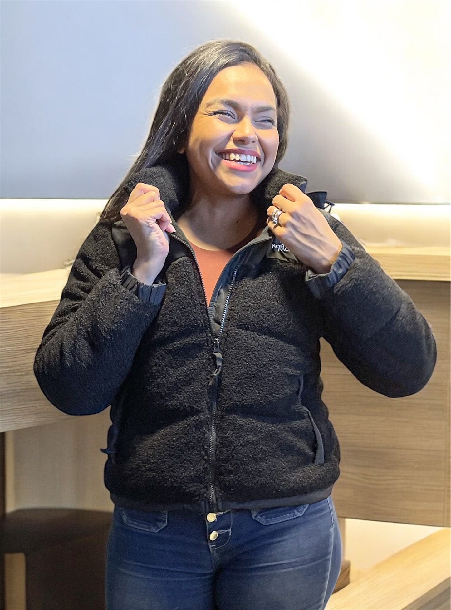 Mujer sonriente usando chaqueta tipo teddy negra, con cierre abierto y textura borreguito visible.